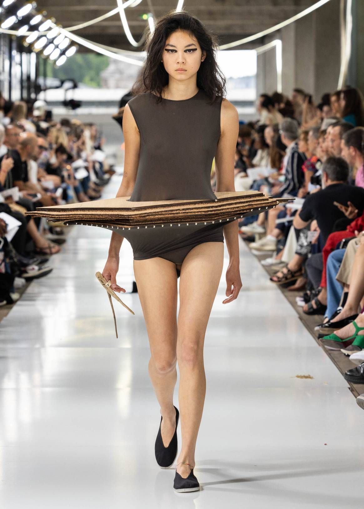 Woman on catwalk wearing a dark-grey body with a horizontal skirt that seems to be made of layers of wood. IFM GRADUATE CLASS 2023.